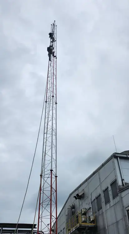 three technicians in safety gear working near the pinnacle of a tall, lattice-style telecommunications tower. 
